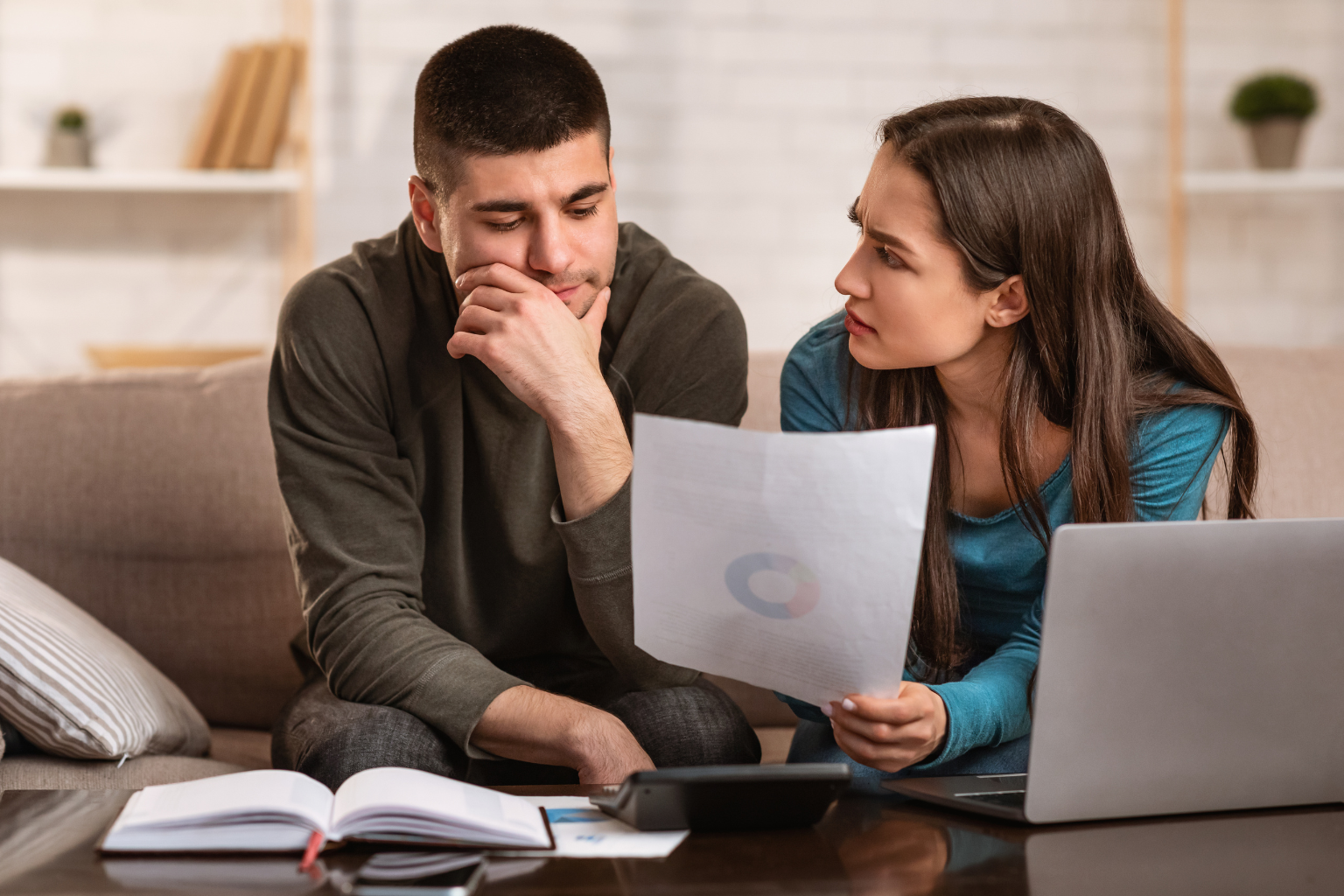 A married couple sits together reviewing financial documents and a laptop, appearing concerned as they discuss their household finances.