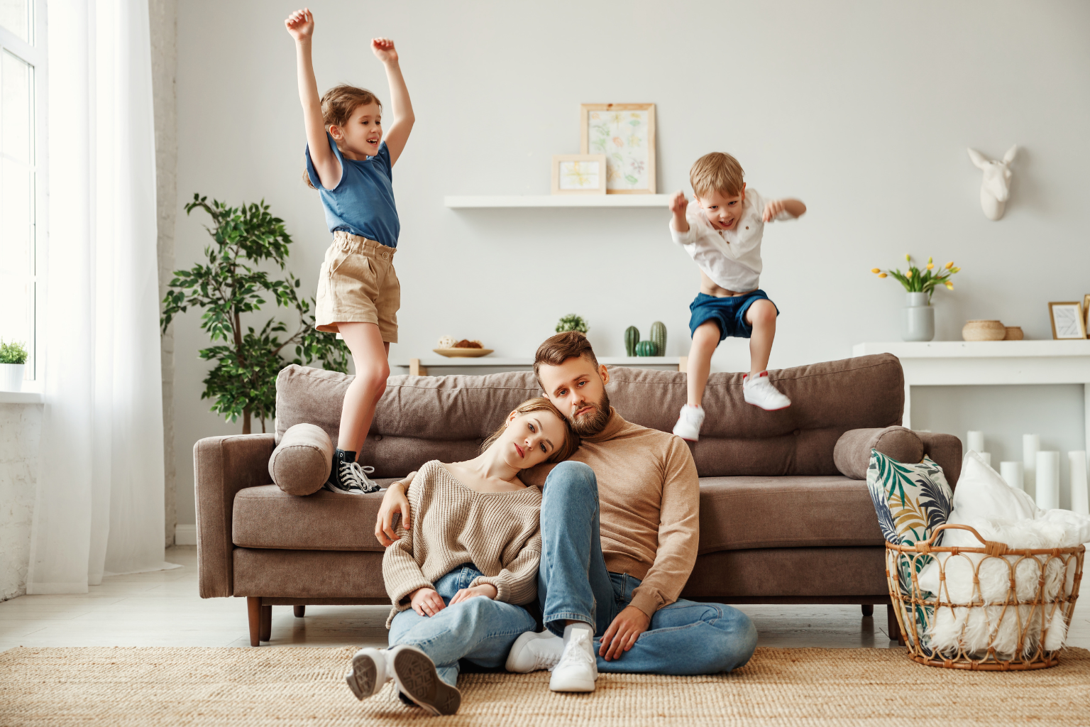 An exhausted mother and father sit closely together on the floor while their two energetic children jump and play on the couch behind them, capturing the joy and exhaustion of family life